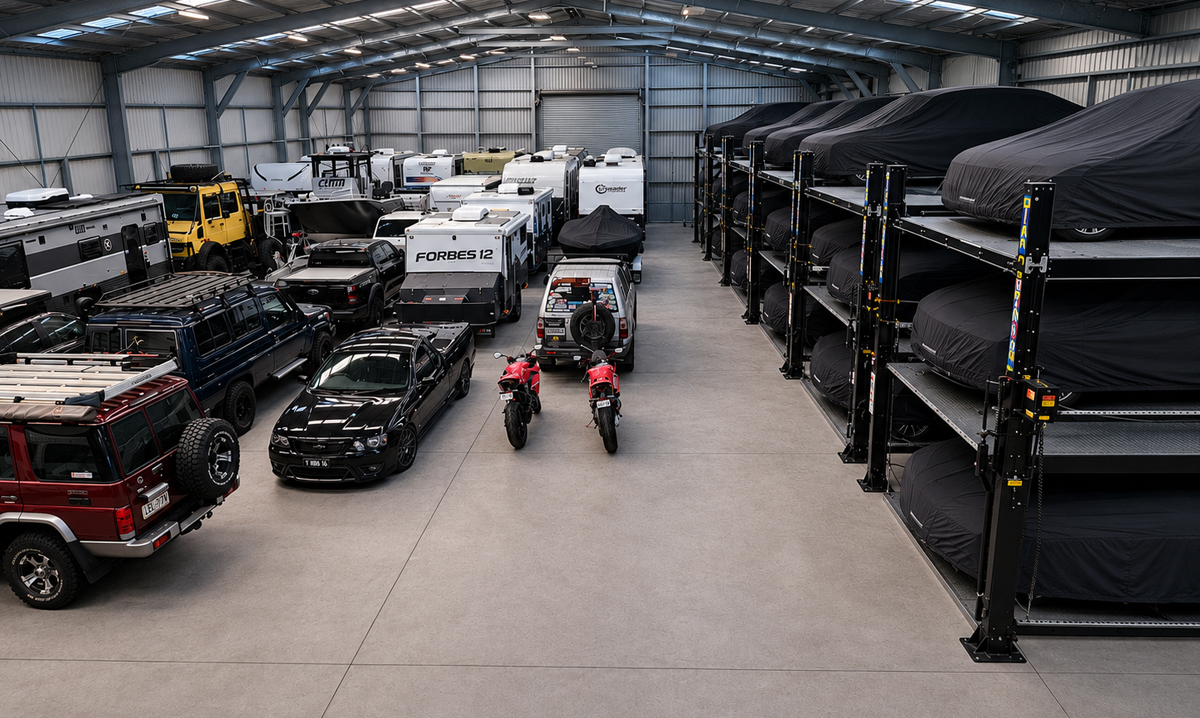 Large indoor storage facility with various vehicles, including cars, motorcycles, SUVs, RVs, and camper vans. Some vehicles are parked on stacked, covered racks, while others are on the ground level.